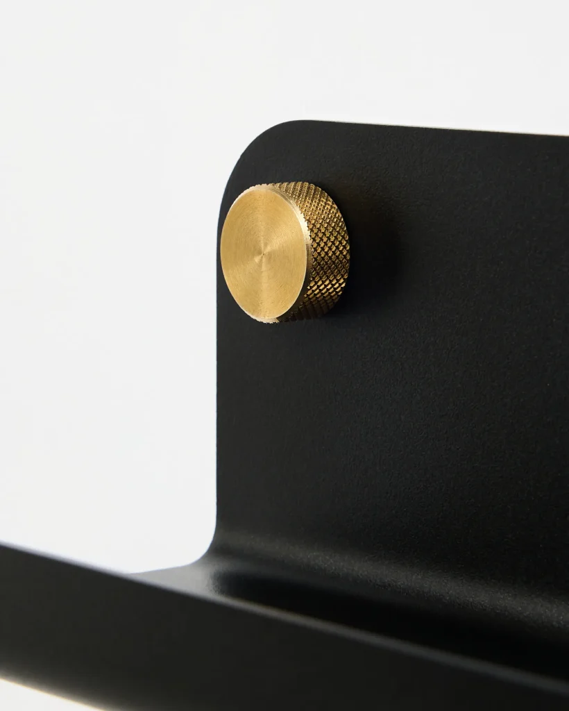 Nahe Ansicht eines WoodUpp-Produkts: eine matte schwarze Metallhalterung oder Regalauflage mit abgerundeter Kante und ein runder, geriffelter Messingknauf in Goldoptik, der als dekorativer Befestigungs- bzw. Stellknopf dient und kontrastreich am Halter angebracht ist, mit feiner zähniger Struktur.
