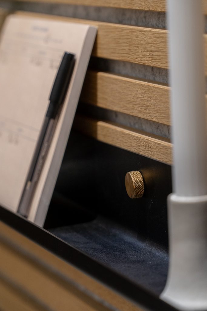 Close-up einer WoodUpp-Wandlösung: horizontale Eichenholzlamellen mit sichtbarer Maserung, davor ein schwarzes Ablagefach mit weichem Samtfutter und ein geriffelter Messingknopf in goldener Optik. Ein Notizblock mit schwarzem Stift steckt in der Nut, mit dezenten, eleganten Akzenten und praktischem Zugriff.