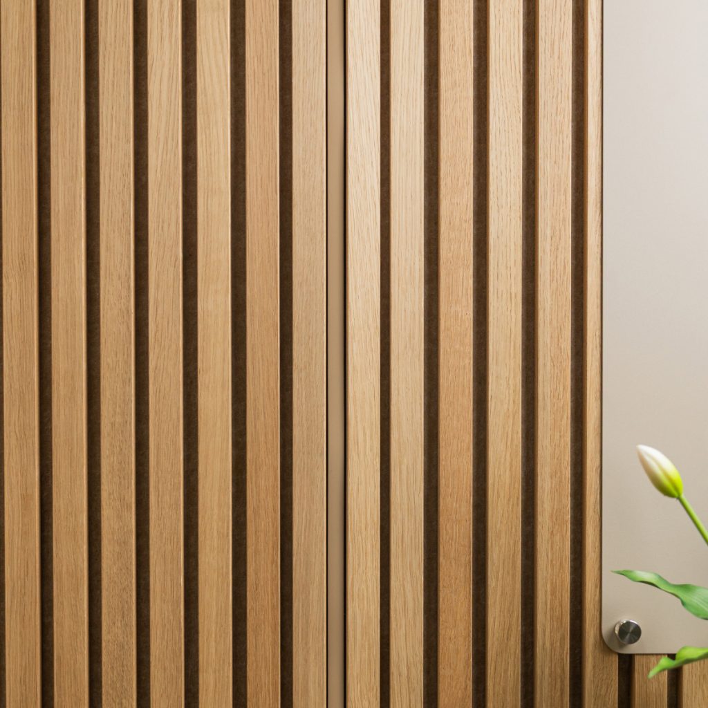 Nahaufnahme einer WoodUpp-Lamellenwand: gleichmäßige, vertikale Lamellenpaneele und feine Holzleisten aus hellem Eichenholz mit schmalen, dunklen Fugen, sichtbarer natürlicher Maserung und mattem Finish, im minimalistischen warmen Design mit betonter vertikaler Linienführung. Rechts eine helle Tafel mit Metallabstandshalter sowie eine einzelne weiße Tulpenknospe mit grünen Blättern.