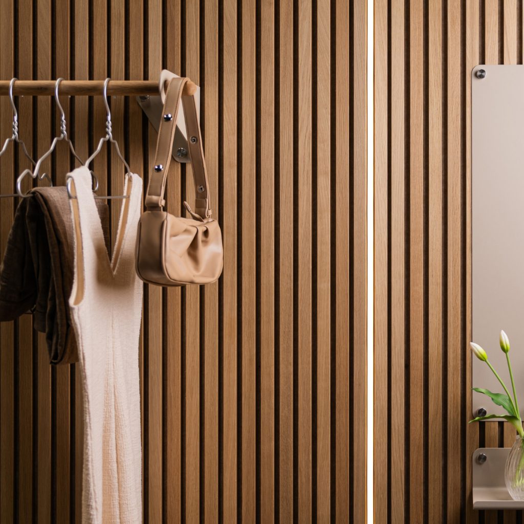 Nahaufnahme einer WoodUpp-Holzlamellenwand aus Eiche mit senkrechten Leisten; daran befestigt eine WoodUpp-Kleiderstange aus Holz mit Metallhalterungen, Haken, hängender Beigetasche und Kleidungsstücken. Rechts montierte WoodUpp-Spiegelhalterung mit kleiner Ablage und Vase mit zarten Knospen. Die warme Holzoberfläche wirkt geölt, Accessoires in Creme- und Beigetönen.