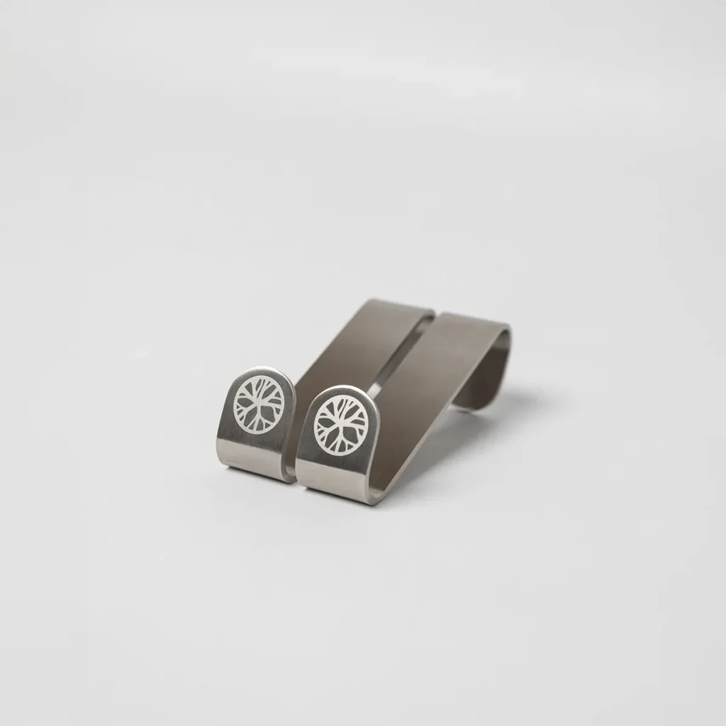 Produktbild von WoodUpp: zwei gebogene silberne Edelstahl-Clips auf weißem Hintergrund. Die kurzen, federnden Metallklammern haben an den vorderen Enden ein eingraviertes WoodUpp-Baumscheiben-Logo in Kreisform. Schlichtes, robustes Design; kompakte Clips zur Befestigung oder Organisation von Kabeln und Gegenständen, mit mattierter Oberfläche und feiner Lasergravur.