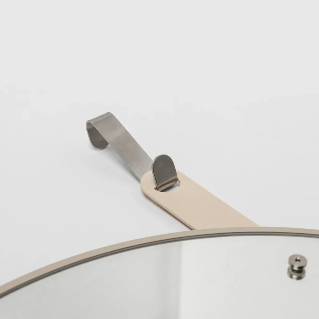 Close-up eines WoodUpp runden Wandspiegels mit dünnem, matt-beigem Metallrahmen. Deutlich zu sehen ist die beigefarbene Aufhängerschlaufe in Lederoptik, die in ein gebürstetes Edelstahl-Hakenstück übergeht, sowie eine kleine sichtbare Metallbefestigungsschraube am Spiegelrand vor hellem Hintergrund. Die Nahaufnahme betont die feine Textur und das elegante, funktionale Design.