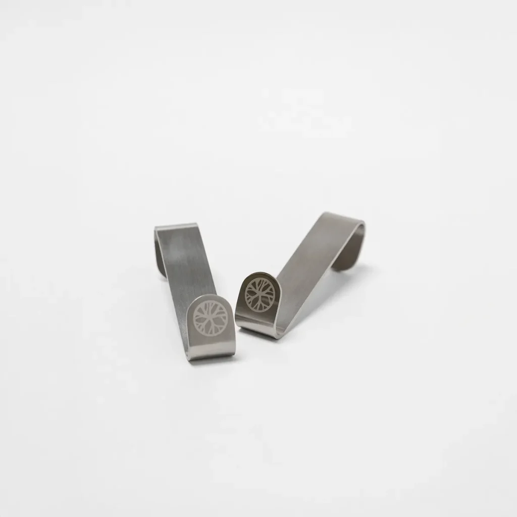 Produktbild von WoodUpp: zwei gebogene, matte Edelstahl-Clips mit offener S‑Form, vorne mit eingraviertem WoodUpp‑Logo (rundes Rad-/Blattmotiv). Die kleinen, flachen Klemmhaken mit fein gebürsteter Oberfläche liegen nebeneinander auf weißem Hintergrund und zeigen schlankes, robustes, minimalistisches Design für Tasche oder Gürtel im Alltag.