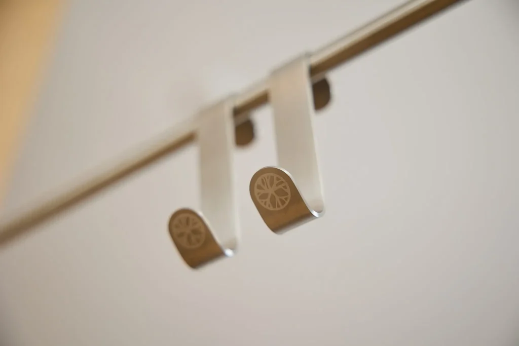 Zwei WoodUpp Türhaken aus gebürstetem Edelstahl hängen über einer metallenen Stange; schlichte U‑Form mit nach unten gebogenem Hakenende. Auf den Hakenenden ist das WoodUpp-Logo (radförmiges zentrales Muster) fein eingraviert. Nahaufnahme, warme Hintergrundbeleuchtung betont matte, silbrige Oberfläche und präzise Kanten.