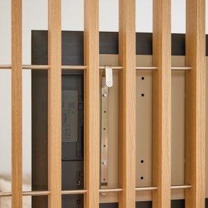 WoodUpp Raumteiler aus hellen Eichen-Lamellen mit quer verlaufenden Metallstäben; dahinter hängt ein Flachbildschirm an einer WoodUpp-Universal-TV-Halterung. Sichtbar sind die stufenlos verstellbare Edelstahl-Aufhängung, Schrauben und die Rückseite des TVs, sauber integrierte Montage für TV und Holzlamellen, stilvolle minimalistische Optik.