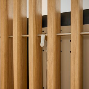 Nahaufnahme einer WoodUpp-Wand mit vertikalen Lamellen aus Eiche in natürlicher Oberfläche, schlanker horizontaler Edelstahlstange, matt gebürstetem WoodUpp-Klemmhaken mit Logo und beige-lackiertem Lochraster-Paneel zur Zubehörbefestigung; warmes Holz, sichtbare Maserung, abgerundete Kanten und präzise, modulare Verarbeitung, mit gleichmäßigem Abstand der Lamellen, feinen Fugen und minimalistischer, neutraler Farbgebung.