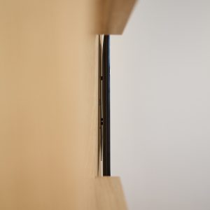 Nahaufnahme einer WoodUpp-Installation: ein schwarzer, schlanker Metall‑TV‑Halter von WoodUpp fixiert einen Flachbildschirm nahezu bündig zwischen zwei massiven, hellen WoodUpp-Holzregalen aus Eiche. Sichtbare Kabelführung, schmale Abstandshalter und präzise Wandbefestigung betonen die flache Montage sowie die elegante, naturbelassene Oberfläche.