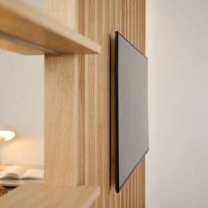 Bild zeigt ein WoodUpp-Element: eine helle, vertikal lamellenförmige Holztrennwand aus Eichenfurnier mit integriertem Regalboden und flacher TV‑Wandhalterung, an der ein schlanker Flachbildschirm mit schwarzem Rand bündig montiert ist; im Vordergrund sichtbar ein Ablagefach mit Lampe und geöffnetem Buch im modernen, minimalistischen Design.