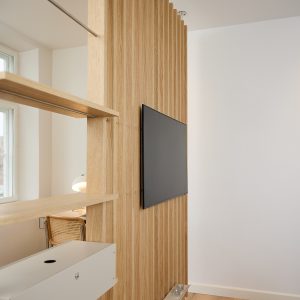 Nahaufnahme einer WoodUpp‑Lamellenwand aus hellem Eichenholz mit schlanken, vertikalen Lamellen als Raumteiler; daran hängt ein flacher Fernseher in Wandhalterung, links sind zwei WoodUpp‑Fachböden/Regalbretter mit Metallaufnahmen befestigt. Die feine Holzmaserung, warme Farbtöne und zwei kleine Kerzengläser auf der Ablage wirken wohnlich.