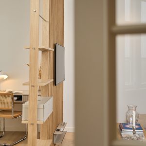 WoodUpp Raumteiler in hellem Eichenholz mit integriertem TV‑Panel, schmaler Metallablage für Fernbedienungen und Kerze sowie einem weißen Aufbewahrungsfach auf einem Regal. Zu sehen sind freistehende Regalböden, ein angrenzender Schreibtisch mit geflochtenem Stuhl, Glasvase auf Beistelltisch und warme Beleuchtung.