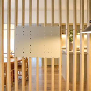 WoodUpp Holzlamellen-Raumteiler aus hellem Eichenholz mit stählernen Querhaltern, daran eine sandfarbene, rechteckige WoodUpp‑Montageplatte aus Metall mit zahlreichen Bohrungen und variabler Lochanordnung für flexible TV‑ oder Regalmontage, sichtbar auf Metallstiften verschraubt und mittig über bodenhohen Lamellen montiert; im Hintergrund Essbereich und offene Küche.