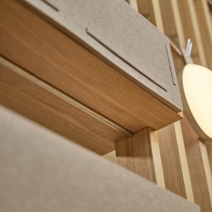 Detailaufnahme einer WoodUpp-Installation: Eichenholzregal mit schlanken Stützen und vertikaler Holzlamellenwand, kombiniert mit hellgrauen Akustikfilzpaneelen von WoodUpp. Im Bild außerdem eine runde Wandlampe; Fokus auf Maserung, Kanten und Verbindung zwischen Holzregal und Filzpaneel. Die präzisen Fugen und der natürliche Eichenfarbton betonen die handwerkliche Qualität der WoodUpp-Holzprodukte.