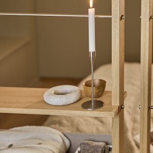 WoodUpp: Nahaufnahme eines offenen WoodUpp-Eichenholz-Regals als Raumteiler mit sichtbaren Metallstreben und Schrauben, einer Ablage mit schlankem Edelstahl-Kerzenhalter (runde Basis) und brennender weißer Stabkerze, einer kleinen WoodUpp-Holzschale sowie einem runden Marmor-Objekt; darunter weiche Decke und gefaltete Textilien, elegante, minimalistische Anmutung in warmem, natürlichem Eichenholzton, zarte Flamme erzeugt stimmungsvolle Beleuchtung.