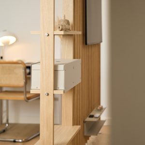 WoodUpp Raumteiler in hellem Eichenholz mit schmalen Regalböden und sichtbaren Befestigungsbolzen; darauf eine hellgraue Metall-Aufbewahrungsbox von WoodUpp, eine kleine Rolle Naturgarn auf dem oberen Brett sowie seitlich eine schlanke Ablagefläche mit Fernbedienungen und einem an der Wand montierten TV-Bildschirm, im Hintergrund Schreibtisch und rattanbesetzter Stuhl.