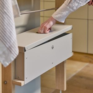 Nahaufnahme einer Person, die mit der Hand den runden Griffdeckel einer weißen WoodUpp Aufbewahrungsbox öffnet; die Box ist an einem hellen Holzgestell unter einer Edelstahl-Kleiderstange befestigt. Sichtbar: runde Grifföffnung, kleine Lüftungsöffnung und Schrauben am matten Deckel. Das minimalistische Design der WoodUpp-Produkte ist gut erkennbar.