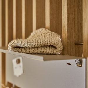 Detailaufnahme eines WoodUpp Garderobenpaneels aus hellen Eichenleisten mit schmalen, vertikalen Lamellen, aufgesetztem grauen WoodUpp Metallfach/Briefkasten mit Magnet-Clip und kleiner Ablage; darauf liegt ein gefalteter, grobgestrickter cremefarbener Wollpullover im warmen, natürlichen Licht, Holzmaserung sichtbar, mattes Finish.