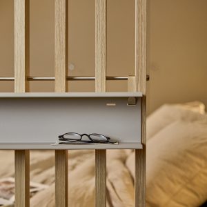 Nahaufnahme eines WoodUpp-Ablageboards: helles Eichenlatten‑Modul mit aufgestecktem, hellgrauem Regalbrett, Metallstange und kleinem Haken. Auf dem matt lackierten Brett liegen eine schwarze Brille und Karten; im Hintergrund unscharf Bett und Kissen. Feine Holzmaserung und dezente goldfarbene Metallhalter betonen das reduzierte Design.