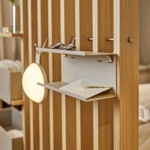 WoodUpp Raumteiler aus hellem Eichenholz mit vertikalen Lamellen und montierter, sandfarbener Metall-Ablage. Zweistufiges, schlankes Regalfach mit Brille oben und Briefumschlag unten, sichtbare Messing-Schraube, kleine Metallhaken und runde Hängeleuchte — dezenter, modularer Ordnungs- und Ablagebereich für Flur, Schlafzimmer oder Büro.