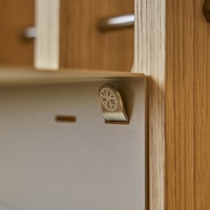 Detailaufnahme eines WoodUpp-Produkts: ein naher Blick auf einen gebürsteten Metall-Klemmhaken mit feiner Radgravur, sicher eingehakt in eine matte, sandfarbene Metallplatte und befestigt am hellen Eichenholzrahmen. Im unscharfen Hintergrund sind weitere Holzlatten und Metallstangen des WoodUpp-Regalsystems sichtbar. Die feine Maserung des Holzes und das minimalistische Design betonen hochwertige Verarbeitung.