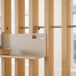 WoodUpp Raumteiler aus schlanken, vertikalen Eichenlamellen mit durchgezogener Metallschiene, an der ein WoodUpp pulverbeschichtetes Hängeregal in hellem Grau befestigt ist; auf dem Regal steht eine kleine, handgedrehte Holzschale. Ein dezenter Metallclip hält das Regal am Stab, sichtbare Eichenmaserung, minimalistisches, skandinavisch inspiriertes Design.