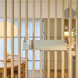 WoodUpp-Holzlamellen als schlanker Raumteiler mit sichtbarer Eichenmaserung, dazu eine montierte WoodUpp-Wandablage in cremefarbenem Metall mit integriertem Kerzenhalter; darauf brennt eine weiße Stabkerze. Im Hintergrund ein runder Esstisch mit Papierleuchte und gedecktem Tisch, skandinavisch inspiriertes Interieur.