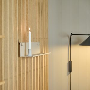 WoodUpp Wandinstallation mit hellen Eichen-Lamellen: eine weiße, minimalistische WoodUpp Wandablage mit flachem Regalboden, sichtbarer Metall-Rückplatte und integriertem Keramik-Kerzenhalter, darauf brennende weiße Stabkerze. Rechts hängt eine schlanke schwarze, schwenkbare Wandleuchte; dezente, neutrale Farbtöne und klare Linien betonen das skandinavische Design.