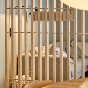 WoodUpp Raumteiler aus hellen Eichenlamellen mit schlanken horizontalen Metallverbindern, die eine filigrane Trennwand bilden. Dahinter ein hellbeiges Sofa mit Kissen und eine dezente Wandleuchte. Natürliche Holzstruktur und klarer, luftiger Raumteiler prägen das gemütliche Wohnambiente. Die vertikalen Lamellen sind gleichmäßig gebohrt und mit dezenten Metallclips befestigt.