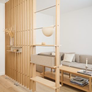 WoodUpp Raumteiler-Regalsystem aus hellem Eichenholz mit senkrechten Lamellen, mehreren Holzablagen, einer herausnehmbaren Filzbox und einer hängenden Halbkugel-Leuchte. Kleine Wandablage mit Vase sichtbar; modulare Metallstangen und Schrauben betonen die funktionale, minimalistische Trennung von Wohn- und Sitzbereich. Auf dem Regal liegen Bücher und ein Kerzenhalter, die warme, natürliche Optik passt zu Parkett