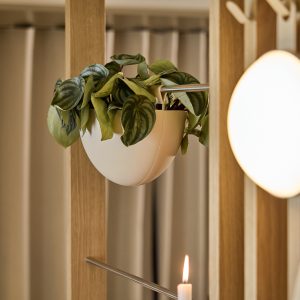 WoodUpp Raumteiler aus hellen Holzlatten mit eingesteckten Metallstangen, an denen ein halbkreisförmiger weißer WoodUpp Pflanzenhalter mit grüner Hängepflanze hängt. Weiter sichtbar: ein schlanker WoodUpp Kerzenhalter mit brennender Kerze und eine runde Wandlampe, alles in warmem, skandinavischem Design für gemütliche Akzente im Innenraum.