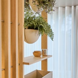 WoodUpp Regal: schlankes, modulares Raumteilersystem aus hellem Eichenholz mit vertikalen Lamellen, zwei offenen Regalböden und einer unteren Aufbewahrungsbox. Daran hängt ein weißer, halbrunder WoodUpp-Pflanzkübel mit üppigen grünen Pflanzen; auf dem Brett stehen kleine Holzschalen und ein glänzender Metallbecher. Das Design wirkt minimalistisch und luftig.