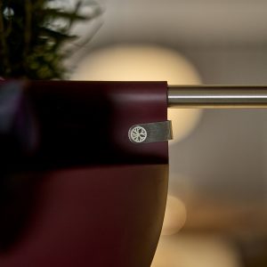 Nahaufnahme eines WoodUpp Hängeblumentopfes in mattem Dunkelrot, befestigt an einer schlanken Edelstahlstange mit flachem, silbernem WoodUpp‑Metallclip, graviert mit einem radialen Rad‑Logo. Der runde Topf zeigt abgerundete Form und matte Oberfläche; oben grünes Blattwerk, im Hintergrund warme, unscharfe Lichter und betont elegantes Hängesystem.