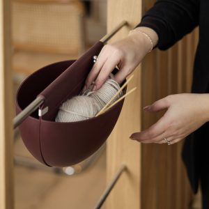 WoodUpp Hängekorb in bordeauxrot, halbrund und aus flexiblem Silikon, an einer Holzstange aufgehängt. Im Korb liegen ein helles Garnknäuel und mehrere glatte Holzstricknadeln; zwei Hände mit Ringen greifen das Garn. Nahaufnahme zeigt praktisches, elegantes Aufbewahrungszubehör, ideal für Wohnzimmer oder Kreativbereich.