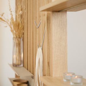WoodUpp Lamellenwand aus hellem Eichenholz mit sichtbarer Maserung, schlanken silbernen Metallhaken in V‑Form sowie schmalen Ablageböden. Auf den WoodUpp-Produkten stehen Kerzen, eine Glasvase mit Trockenblumen und kleine Schalen; an einem Haken hängt ein cremefarbener Stoffbeutel – elegante, minimalistische Optik, wohnlich.