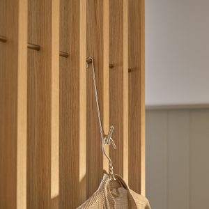 Detailaufnahme eines WoodUpp Eichenlamellen-Paneels im Flur: warmes, naturgeöltes Eichenholz mit schmalem Lamellenraster, feiner Maserung und mattem Finish. Eingelassene zylindrische Messinghaken bilden eine dezente Hakenleiste. An einem Haken hängt ein schlanker silberner Metallbügel mit beigem Baumwollnetzbeutel. Minimalistisch, funktional und handwerklich gefertigt für ein elegantes, einladendes Ambiente.