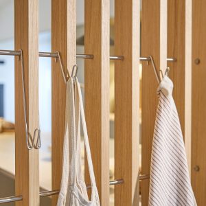 WoodUpp Raumteiler aus hellem Eichenholz mit schlanken vertikalen Lamellen, eingesetzten Edelstahlstangen und abnehmbaren Edelstahlhaken. An den Haken hängen ein naturfarbener Baumwollbeutel und ein weiß-rot gestreiftes Küchentuch. Klare Maserung, minimalistische Optik und stabile Verarbeitung betonen skandinavisches Design. Ideal als Garderobe, Handtuchhalter oder Raumteiler.