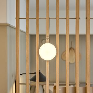 WoodUpp Lamellenwand aus hellem Eichenholz als Raumteiler mit sichtbarer Metallstange, an der eine WoodUpp Hängeleuchte mit rundem, opalfarbenem Diffusor hängt und warmes, gleichmäßiges Licht abstrahlt. Davor ein schwarzer Leselampenarm; im Hintergrund dezente Wandverkleidung und natürliche, organische Dekoration, skandinavisch inspiriertes Interieur in ruhigen Farbtönen.