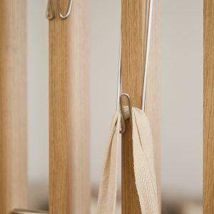 WoodUpp zeigt eine Nahaufnahme einer Garderobenlösung: schmale, senkrechte Eichenholzlamellen mit sichtbarer Maserung und warmem Ton sowie schlanke, verchromte Metallhaken von WoodUpp mit glänzender Oberfläche. An einem Haken hängt ein naturfarbener, grob gewebter Stoffriemen, und helle, ruhige Farbstimmung.