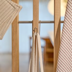 WoodUpp Wandpaneel aus hellem Eichenholz mit sichtbarer Maserung, quer eingesetzter, polierter Edelstahlstange und schlanken Metallhaken. Schlichte, skandinavisch inspirierte Optik. An den Haken hängen ein beigefarbener Topflappen mit feinen Dunkelstreifen, ein rot-weiß gestreiftes Geschirrtuch sowie mehrere naturfarbene, gewebte Baumwoll-Trägerbänder, praktisch und dekorativ im Küchenbereich.