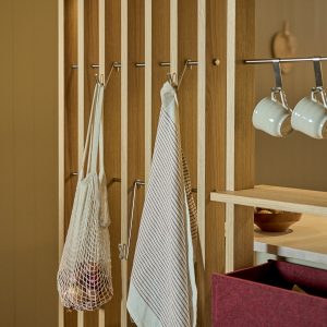 WoodUpp Holzlamellen-Trennwand mit schlanken Eichenstäben, integrierter Metallhängeschiene und mehreren Edelstahlhaken, an denen zwei weiße Kaffeetassen hängen. Zusätzliche Haken halten ein Netz-Einkaufsbeutel und ein gestreiftes Küchentuch; seitlich eine Holzablage mit Schale und ein roter Filz-Aufbewahrungsbehälter. Das modulare, minimalistische Design betont natürliche Holzmaserung und vielseitige Aufbewahrungsmöglichkeiten.