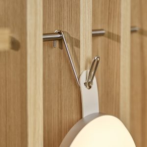 Nahaufnahme eines WoodUpp Eichen-Paneels mit schmalen Lamellen, an dem ein verchromter Metallhaken befestigt ist. An dem Haken hängt eine runde, weiße WoodUpp-Lampe mit Schlaufe. Klare, warme Holzmaserung, matte Metalloberfläche und weiches Licht der Lampe sichtbar. Betont funktionales, minimalistisches Design und filigrane Verarbeitung.