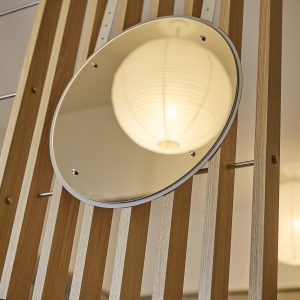 WoodUpp Holzlamellen-Trennwand aus hellem Eichenholz mit schmalen, vertikalen Lamellen und quer eingesetzten, mattierten Metallstangen; daran montiert ein runder WoodUpp-Wandspiegel mit Aluminiumrahmen und polierten Befestigungsbolzen. Im Spiegel spiegelt sich die warme, runde Lichtreflexion einer Papierlaternen‑Hängelampe. Der Spiegel hängt leicht schräg und betont die dezente, skandinavisch-minimalistische Ästhetik der WoodUpp-Module.