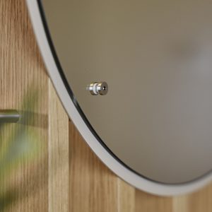 Nahaufnahme eines WoodUpp-Interieurs: runder Wandspiegel mit schmalem weißen Rahmen und dunkler Einfassung, montiert an einem glänzenden Edelstahl-Abstandshalter auf hellen WoodUpp-Holzlamellen in Eichenoptik. Unscharfer Pflanzenvordergrund links, der Schraubenkopf des Abstandshalters zeigt minimalistisches, hochwertiges Design und schafft warme, moderne Badezimmer- oder Flur-Atmosphäre.