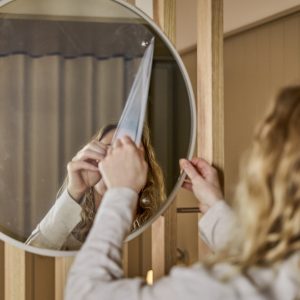 Eine Person zieht die Schutzfolie von einem runden Spiegel der Marke WoodUpp ab, montiert in einem schmalen, hellen Holzrahmen vor vertikalen WoodUpp-Holzlatten. Zu sehen sind der runde Wandspiegel mit dünnem Rahmen, die modulare, helle Holzpaneel-Befestigung und das minimalistische Design von WoodUpp im warmen Raumambiente.