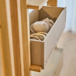 Nahaufnahme eines WoodUpp-Standregals aus hellem Eichenholz mit herausziehbarer WoodUpp-Filzbox in Grau. In der Box liegen beige Wollknäuel und mehrere Holzstickringe. Das minimalistische Möbel zeigt feine Holzleisten; die Box sitzt auf einem schmalen Holzgriff in warmem, neutralem Ambiente für Strick- und Bastelutensilien.