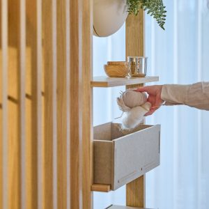 WoodUpp elegantes Raumteiler-Regal aus hellem Eichenholz mit schlanken Leisten, offener Ablage mit kleiner Holzschale und Metallbecher sowie einer filzbezogenen Aufbewahrungsbox; daran ein hängender Pflanztopf. Eine Hand legt Garnbälle in die Box, Szene zeigt funktionale Ordnung und natürliches, warmes, minimalistisches Design.