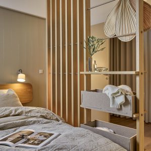 WoodUpp Raumteiler/Offenes Garderobensystem aus hellem Eichenholz mit senkrechten Latten, integrierter Metall-Kleiderstange, zwei grauen Filz-Boxschubladen sowie offenen Holzregalen. Auf den Regalen: Vase mit Zweigen und Dekorgegenständen, im Fach eine weiße Strickjacke; im Vordergrund Bett mit Bettdecke und geöffnetem Magazin und warmer Lichtstimmung.
