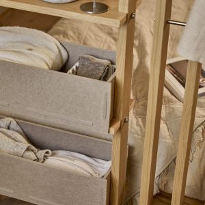 Detailaufnahme eines WoodUpp Eichenrahmen-Regals mit zwei beigen Filz-Aufbewahrungsboxen, gefüllt mit gefalteten Strickwaren und Socken. Die schmale, helle Holzstruktur zeigt sichtbare Metallbeschläge und kleine Ablageflächen; die weichen Filzschubladen mit Steckverbindungen betonen skandinavisch-natürliches Design, warmen Eichenholzton, feine Nähte und dezenten, minimalistischen Stil.