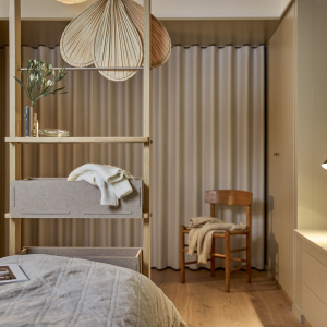 Dieses Schlafzimmer zeigt ein freistehendes WoodUpp Regal aus heller Eiche, das als stilvoller Raumtrenner fungiert. Es verfügt über integrierte graue Filzaufbewahrungsboxen mit Strickwaren und Holzböden mit Dekoration wie Vasen und großen Fächern. Ein Bett und ein Holzstuhl im Hintergrund runden das gemütliche skandinavische Design ab.