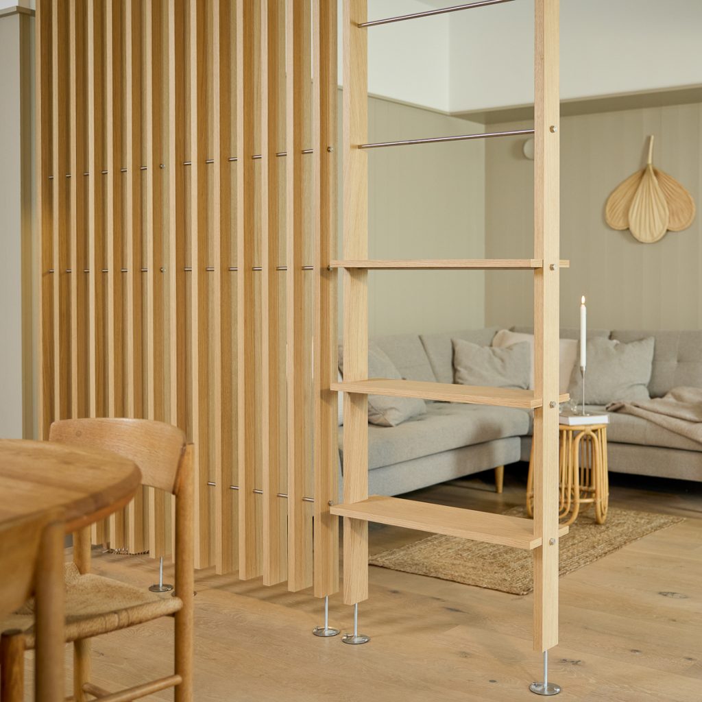 WoodUpp Lamellen‑Trennwand aus hellem Eichenholz neben einem schmalen WoodUpp Regal-/Leiter‑Element mit offenen Ablagen, Metallstangen und verstellbaren Fußplatten, als Raumteiler zwischen Essbereich und Sofaecke, sichtbare Kerze und beiges Sofa im Hintergrund, mit warmem Holzton, schlanken vertikalen Lamellen, offenem, minimalistischem skandinavischem Design und gemütlicher Atmosphäre.