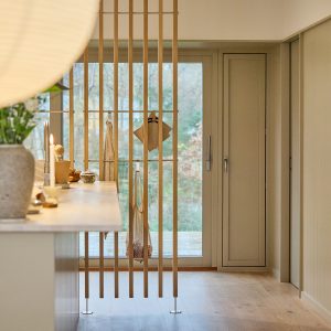 WoodUpp-Raumteiler aus schlanken, senkrechten Holzlatten mit sichtbaren Metallfüßen und Querverbindungen, separiert Küche und Flur. An der Konstruktion hängen WoodUpp-Kleiderhaken und eine horizontale Stange mit Netzbeutel und Topflappen. Im Vordergrund eine helle Küchenarbeitsplatte mit Kerze und Vase, sanftes Tageslicht, helle Holzoberfläche und skandinavisch-minimalistische Optik.